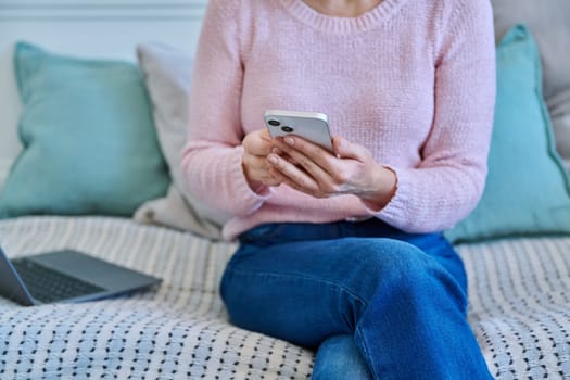 Close-up of smartphone in hands of woman sitting on couch at home. Modern technologies, mobile applications apps for leisure, communication, entertainment, work, learning, reading, shopping concept