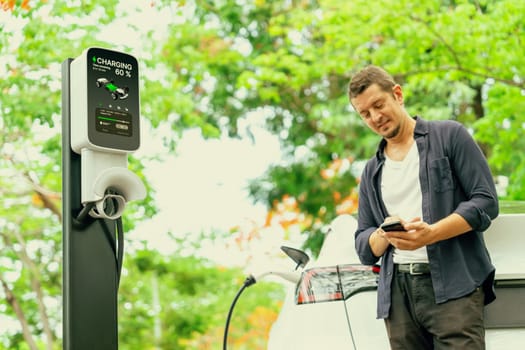 Man using smartphone online banking application to pay for electric car battery charging from EV charging station during autumn vacation holiday trip at national park or autumnal forest. Exalt