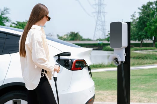 Young woman recharge EV car battery at charging station connected to power grid tower electrical industrial facility as electrical industry for eco friendly vehicle utilization. Expedient