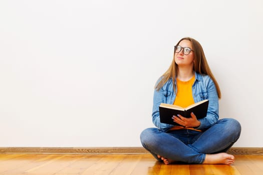 Thoughtful young woman in casual attire sitting on the floor with a book, looking upwards as if in contemplation or inspiration.