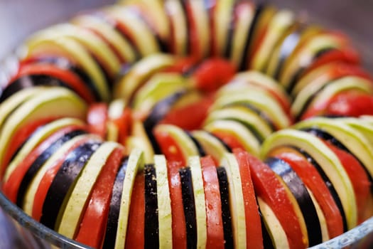 Sliced zucchini, eggplant, and tomato arranged in a spiral in a glass baking dish.