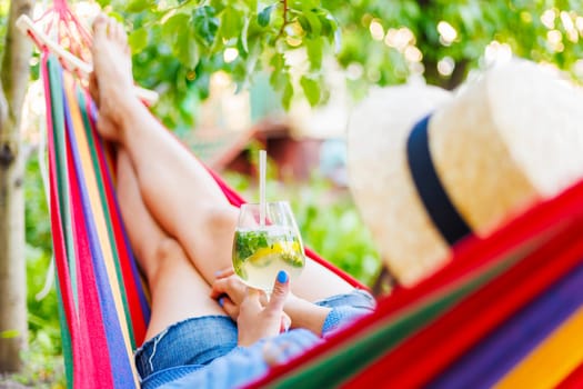 Close-up of bare feet and a chilled mojito in hand, as someone relaxes in a multicolored hammock, symbolizing summer leisure.
