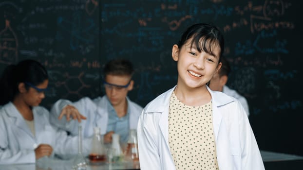 Cute asian scientist smile to camera while diverse group doing experiment. Young happy schoolgirl standing at blackboard with chemical theory written while wearing lab coat at laboratory. Edification.