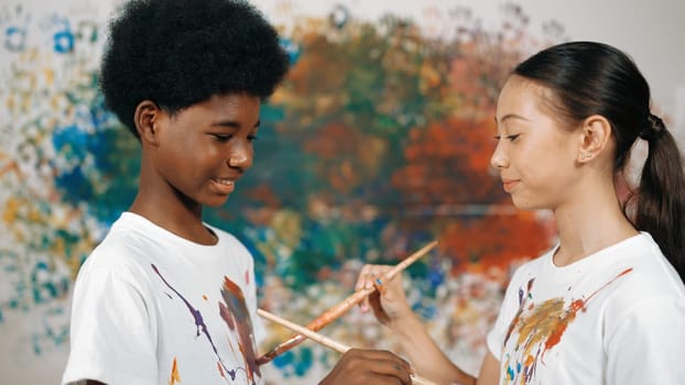 Smiling diverse children using paint brush painted color on each other white shirt shirt at colorful stained wall in art lesson. Represent exchanging experience, learning each other. Edification.
