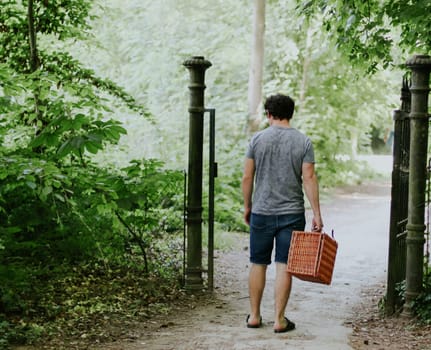 A young caucasian guy with curly brown hair in shorts and a T-shirt from the back walks to the main entrance of a public park with a wicker basket in his hands for a solitary picnic, side view close-up. PARKS and REC concept, picnic, outdoors, relaxing in the park.