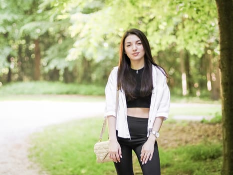 Portrait of one young beautiful Caucasian brunette girl with long fluffy hair in black shorts and a white shirt stands and looks at the camera with a smile in the forest on a spring day, close-up side view.