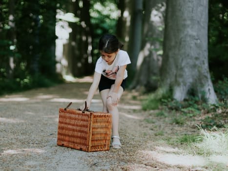 A beautiful Caucasian little girl of six years old takes a wicker basket by its leather handle while standing on a path among the trees in a public park to have a picnic, close-up side view. Concept of picnic time, outdoor recreation.
