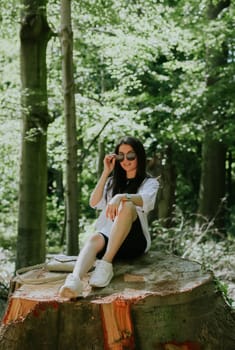 One beautiful young Caucasian brunette girl in sunglasses and stylish clothes poses while sitting on a large sawn stump in the park on a sunny spring day, close-up side view.
