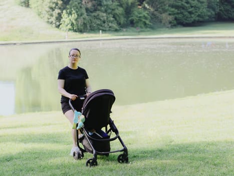 One young beautiful Caucasian girl-mother in black clothes walks with a child in a black stroller along the lawn in a park near a lake on a clear sunny and spring day, close-up side view.