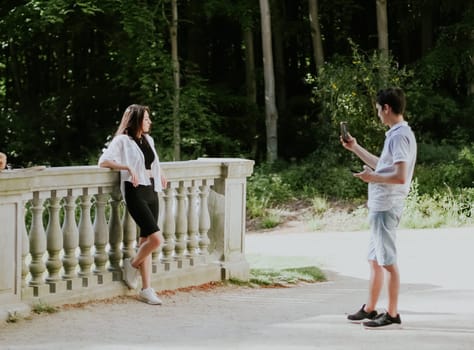 Handsome young caucasian guy photographing a girl leaning on a concrete vintage railing in a park, close-up side view.PARKS concept and REC concept, family vacation, using technology.