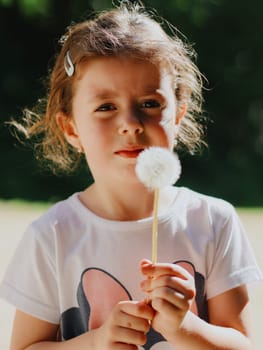 Portrait of one small beautiful Caucasian girl holding a dandelion in her hands and looking at the camera, standing in the park on a spring day, close-up side view with depth of field.