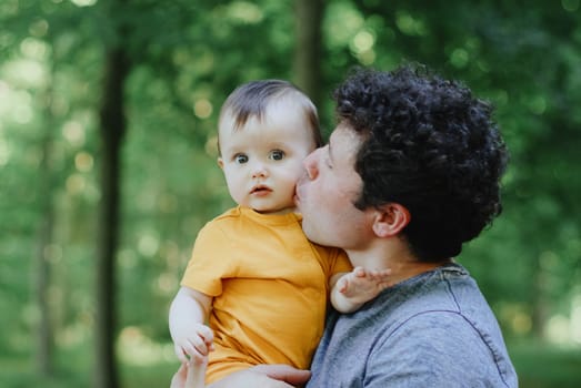 Portrait of one young handsome man father with curly hair holding his little girl daughter in his arms and tenderly kissing her on the cheek while standing in the park on a spring day, close-up side view with depth of field.