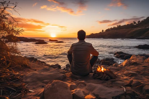 A man admires the sunset on the shore of the bay next to the fire.