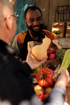 Fresh products for elderly client at organic food market, man talking to vendor and shopping at local farmers market. Friendly farmer standing behind fruit and vegetable stall and serving customer.