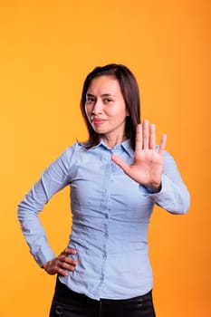 Serious asian woman raising palm to advertise stop gesture in studio over yellow background, expressing denial and refusal. Negative young adult showing rejection gesture, doing rejection symbol