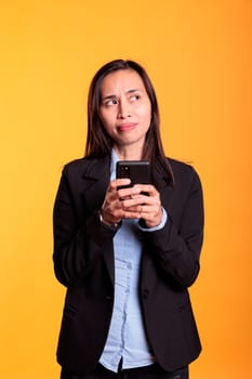 Pensive asian model typing message on smartphone, discussing with remote coworker in studio over yellow background. Cheerful woman in formal suit browsing information on internet, working at project