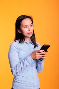 Pensive woman typing message on mobile phone, discussing with remote friend in studio over yellow background. Cheerful filipino young adult browsing on internet, searching information for project
