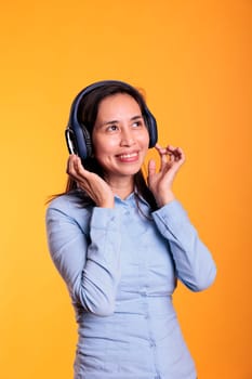 Cheerful filipino woman listening music and dancing, enjoying free time in studio over yellow background. Attractive model wearing headphones showing funny dance moves. Entertainment concept