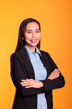 Positive brunette woman standing in arm crossed posing over yellow background, smiling at camera during studio shoot. Smiling filipino adult with cheerful expression enjoying break time