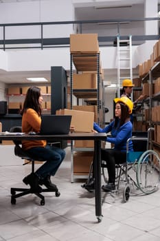 Asian postal service worker in wheelchair and operator maintaining parcel delivery. Storehouse order packer with disability giving cardboard box to distribution manager in storage room