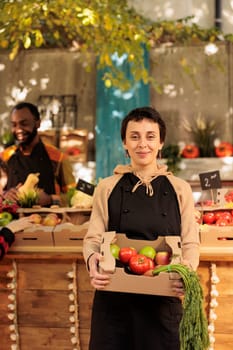 Young happy woman fruit and vegetable market stall owner holding fresh organic produce box in hands and smiling at camera. Female farmer selling healthy local food from roadside stand