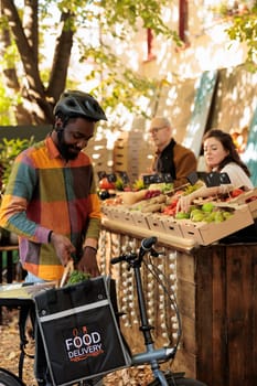 Organic produce delivery services worker picking up food order at local farmers market stand, delivering fresh organic vegetables and fruits. Courier taking farm produce on bicycle.