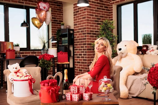 Smiling girlfriend in black dress posing with romantic surprise, receiving luxury presents from boyfriend during valentine s day. Attractive woman sitting in living room filled with red roses and teady bear