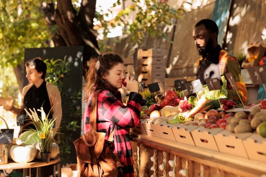 Young woman enjoying shopping at local farmers market on sunny autumn day. Female smelling fresh ripe apple with closed eyes while buying organic food fruits and vegetables, visiting harvest festival