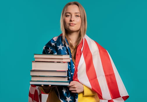 American woman student holds stack of university books from college library on blue background. Happy girl smiles, she is happy to graduate in USA, education abroad concept