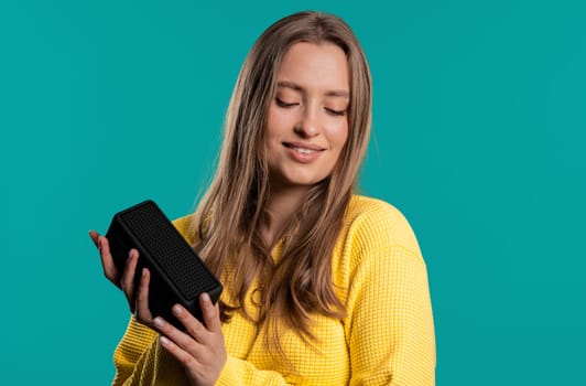 Woman dancing, enjoying on blue studio background. Girl moves to rhythm of music. Young teenager listening to music by wireless portable speaker - modern sound system.