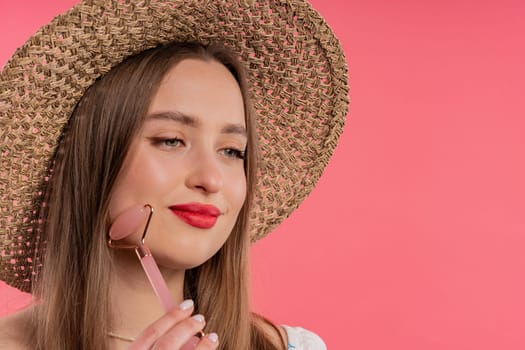 Young woman with rose quartz stone roller on pink background. Facial self care, beauty rituals, cosmetology, anti aging and anti-wrinkle treatment. Copy space. High quality photo