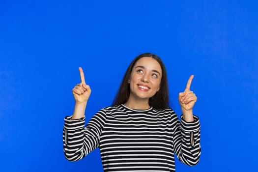 Caucasian smiling woman showing thumbs up pointing overhead empty place, advertising area for commercial text copy space for goods promotion advertisement. Girl isolated on blue background. Copy-space
