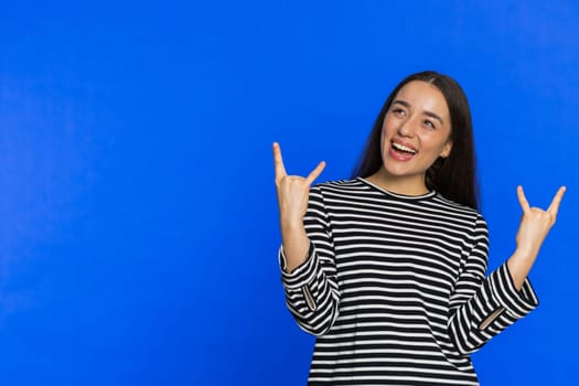 Overjoyed happy Caucasian woman in striped blouse showing rock n roll gesture by hands, cool sign, shouting yeah with crazy expression, dancing, rejoicing victory win success. Girl on blue background