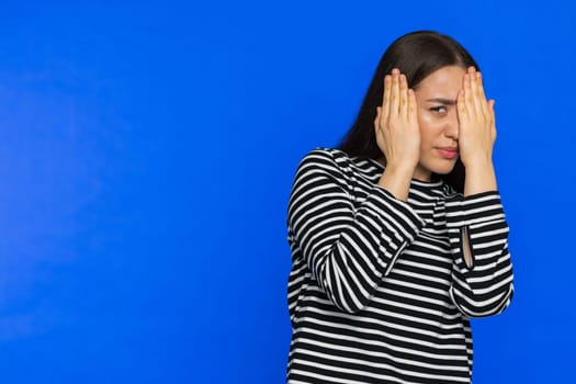 Don't want to look at this, awful. Afraid shocked woman in striped blouse closing eyes with hand showing stop gesture, confused shy scared reject to watch. Girl isolated on blue background. Copy-space