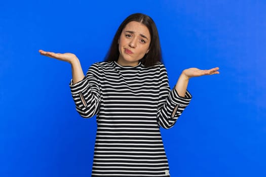 No idea, I don't know answer. Puzzled clueless uncertain Caucasian young woman in striped blouse raising hands in helpless gesture, embarrassed confused by difficult question. Girl on blue background