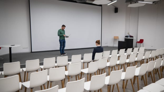 A red-haired Caucasian businesswoman sits in the front row of an empty conference room. Bearded man giving a lecture