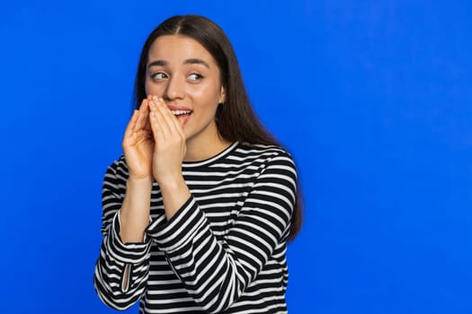 Keep my secrets, silence. Young woman whisper news rumors holding hands near mouth, share gossip, quiet. Girl telling interesting confidential story isolated on blue studio background. Copy-space