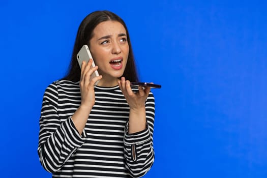 Irritated nervous stressed woman talking screaming on two mobile phones having conversation conflict quarrel complaint dispute discuss solve problem. Young girl isolated on blue background. Copy-space