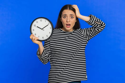 Caucasian woman with anxiety checking time on clock, running late to work, being in delay, deadline. Shocked girl looking at hour, minutes, worrying to be punctual isolated on studio blue background