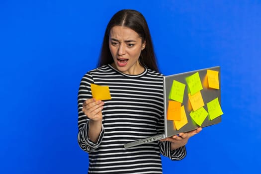 Exhausted freelancer Caucasian woman with pasted sticker notes use laptop computer having concentration problem creative crisis. Nervous young girl confused by big routine work loading blue background