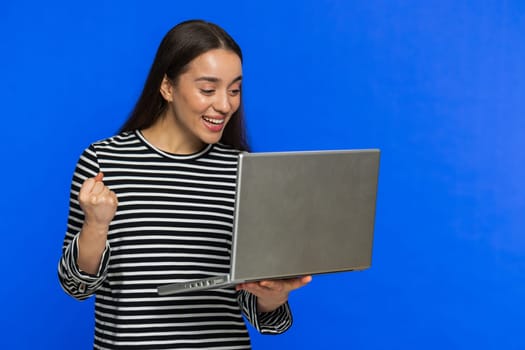 Happy excited Caucasian woman typing on laptop, working on project, enjoying results, winning lottery game celebrating success, online shopping victory good email news. Pretty girl on blue background