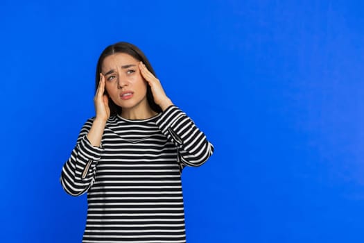 Exhausted young woman girl rubbing temples to cure headache problem, suffering from tension, migraine stress, grimacing in pain, high blood pressure isolated alone on blue wall background. Copy-space