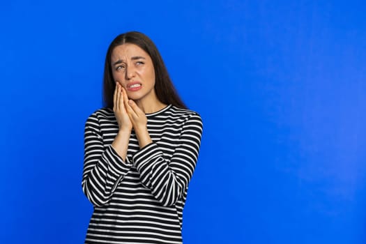 Dental problems. Young woman touching cheek, closing eyes with expression of terrible suffer from painful toothache sensitive teeth cavities. Pretty girl isolated on blue studio background. Copy-space