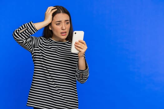 Upset sad Caucasian woman use smartphone typing browsing, play game, loses, surprised by sudden lottery results bad fortune loss fail unlucky news. Pretty girl isolated on blue background. Copy-space