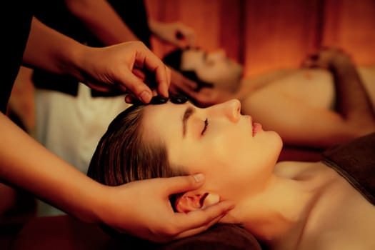 Caucasian couple enjoying relaxing anti-stress head massage with hot stone and pampering facial beauty skin recreation leisure in warm candle light ambient salon spa in resort or hotel. Quiescent