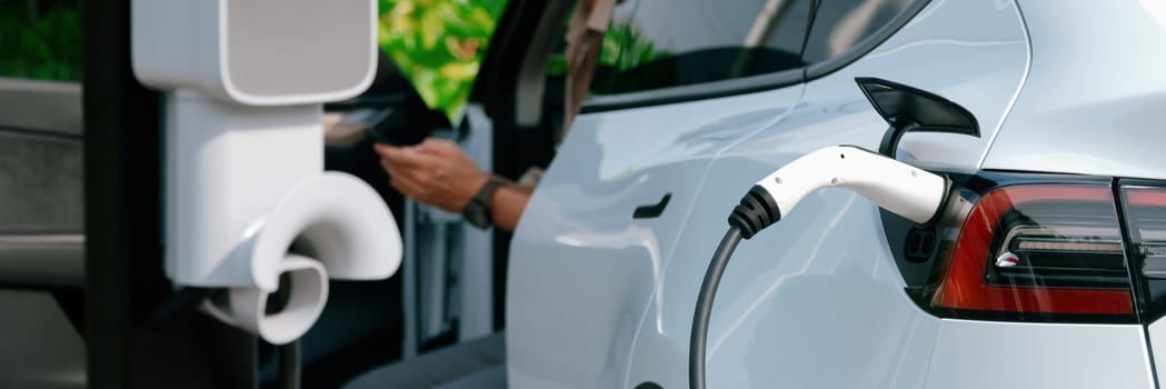 Young man recharge electric car's battery from charging station in outdoor green city park. Rechargeable EV car for sustainable environmental friendly urban travel. Panorama Expedient