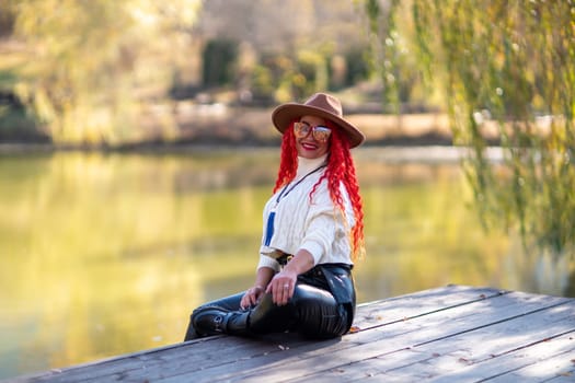 Autumn lake woman. In autumn, she sits by the pond on a wooden pier and admires nature with red hair and a hat. Tourism concept, weekend outside the city