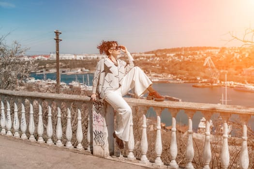 Woman walks around the city, lifestyle. A young beautiful woman in white trousers and a sweater sits on a white fence with balusters and overlooks the sea bay and the city