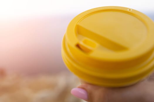 Hand holding Yellow cup with lid, coffee against a backdrop of a blue sky and sea. Illustrating cup and beverage.