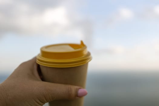 Hand holding Yellow cup with lid, coffee against a backdrop of a blue sky and sea. Illustrating cup and beverage.